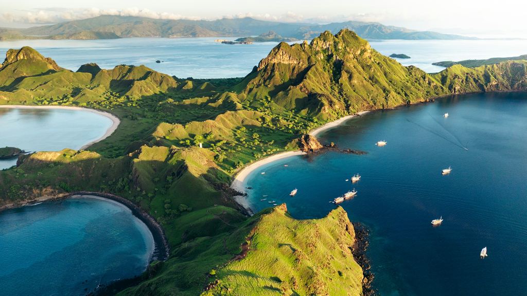 Aerial view of a Komodo National Park island with three curved bays and liveaboard boats at anchor offshore