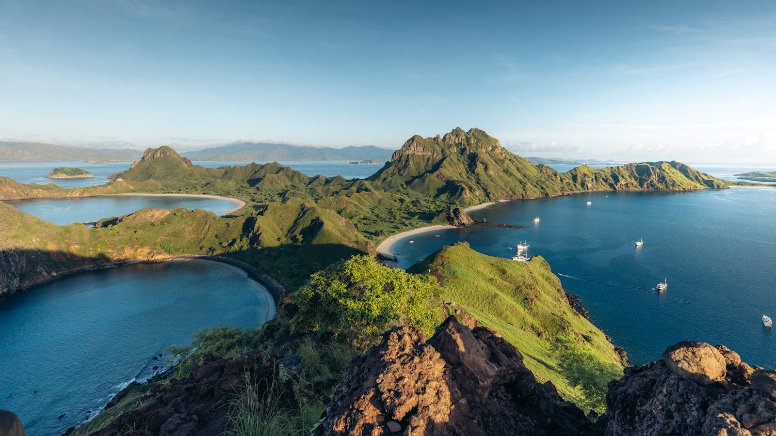 Bays, ridges and anchored boats in Komodo National Park photographed from a summit viewpoint, with rock and scrub in the foreground