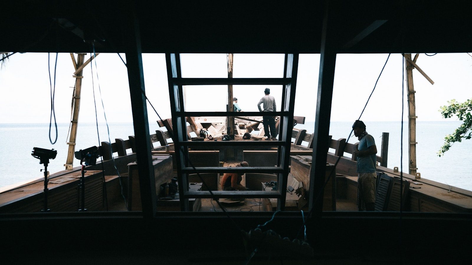 Silhouetted workers constructing a wooden boat by the sea