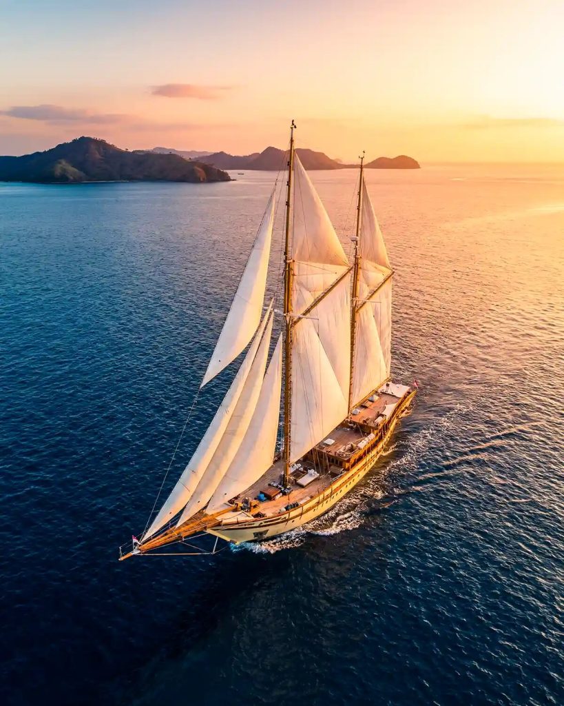 A phinisi under full sail on open water in golden light, seen from above