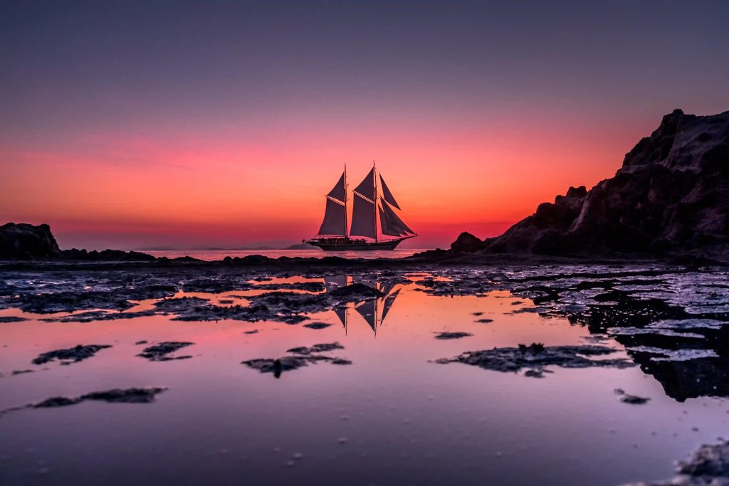 A phinisi silhouetted against a pink and violet sunset, mirrored in the shallows