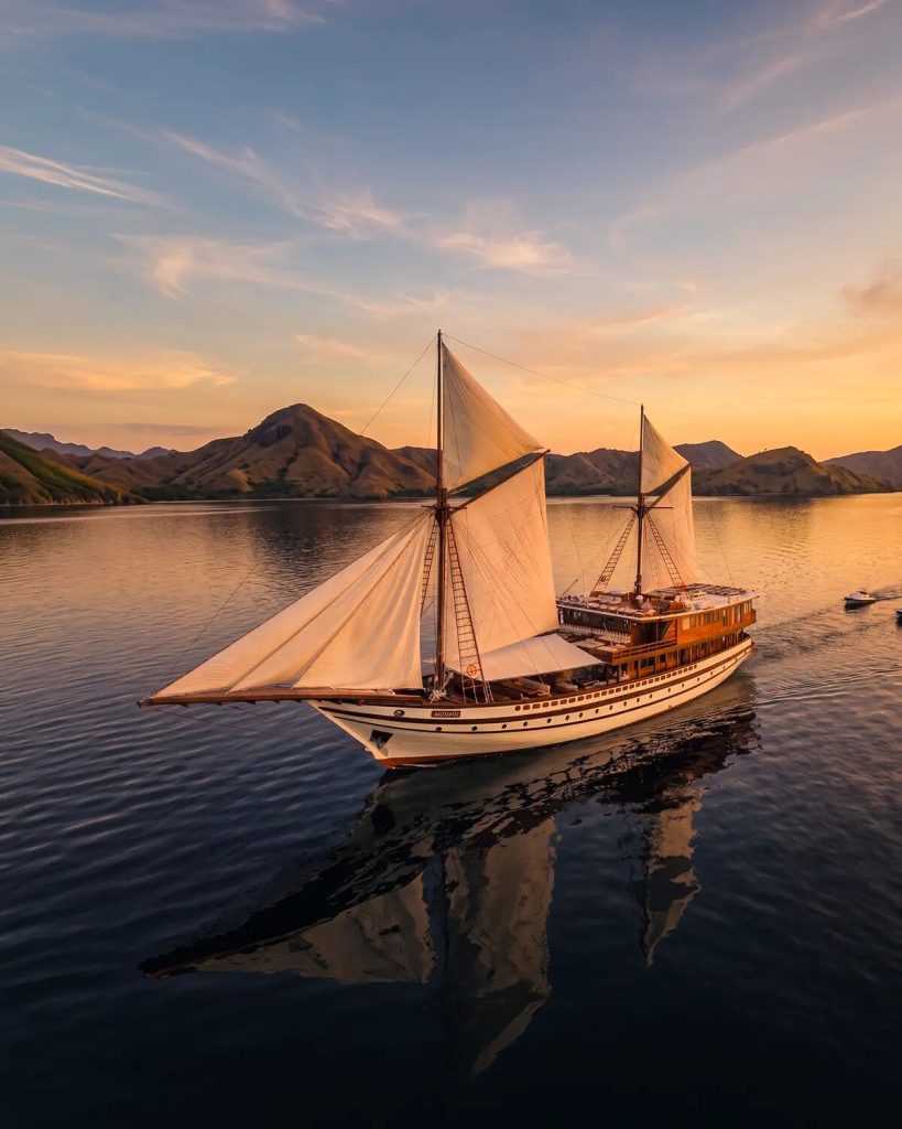 A phinisi under full sail on a still sea at sunset, with dry hills behind