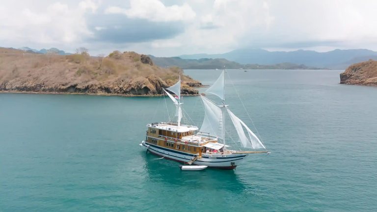 A liveaboard under sail off a dry island in pale green water
