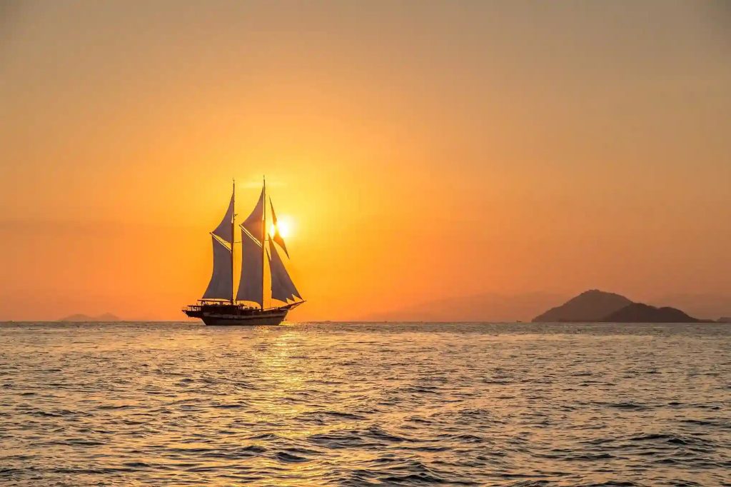 A phinisi under sail silhouetted against an orange sunset on open water