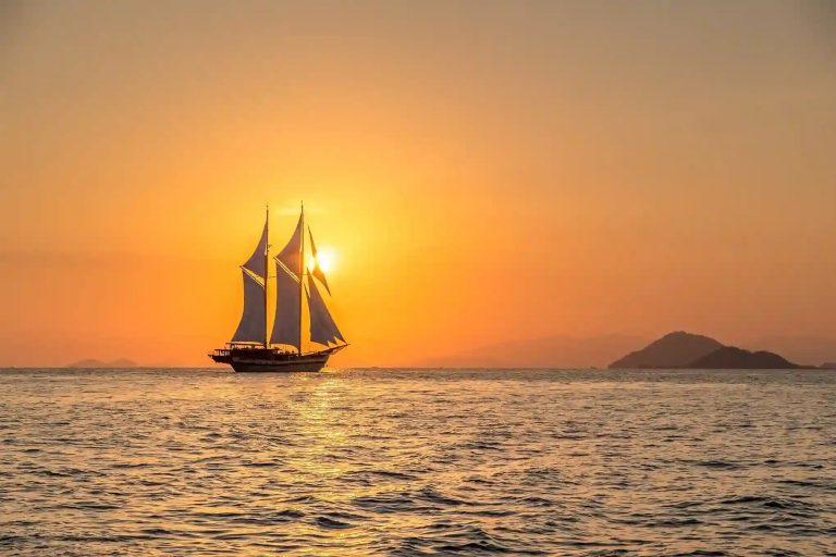 A phinisi under sail silhouetted against an orange sunset on open water