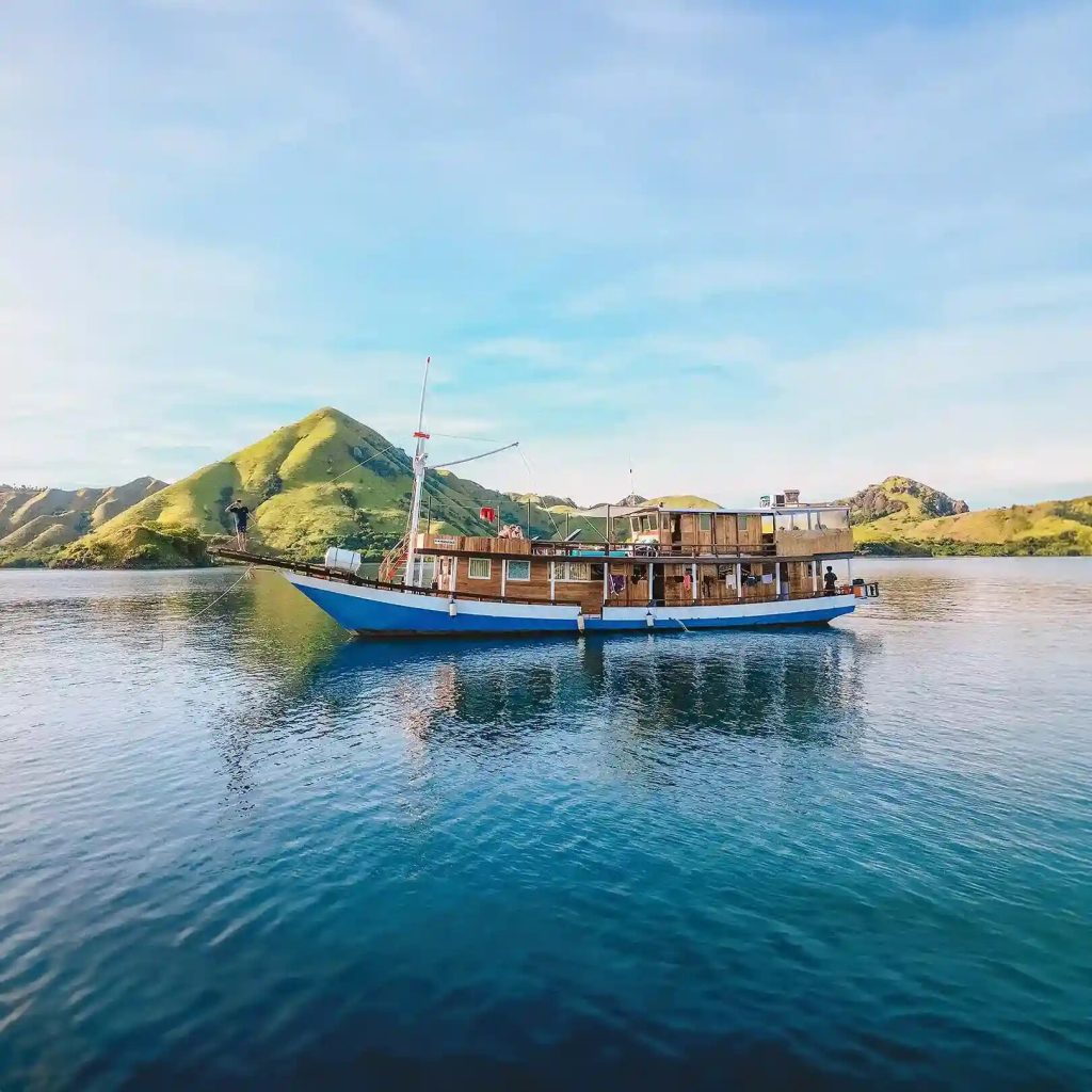 A blue-hulled wooden passenger boat moored below green hills near Labuan Bajo