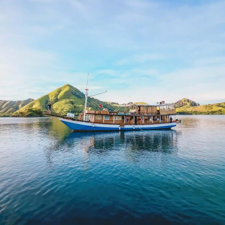 A blue-hulled wooden passenger boat moored below green hills near Labuan Bajo